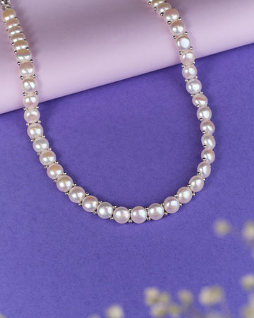 The Smoth Ripple Pearl Necklace by Chandrani Pearls is elegantly displayed with silver accents on a purple and lilac surface.