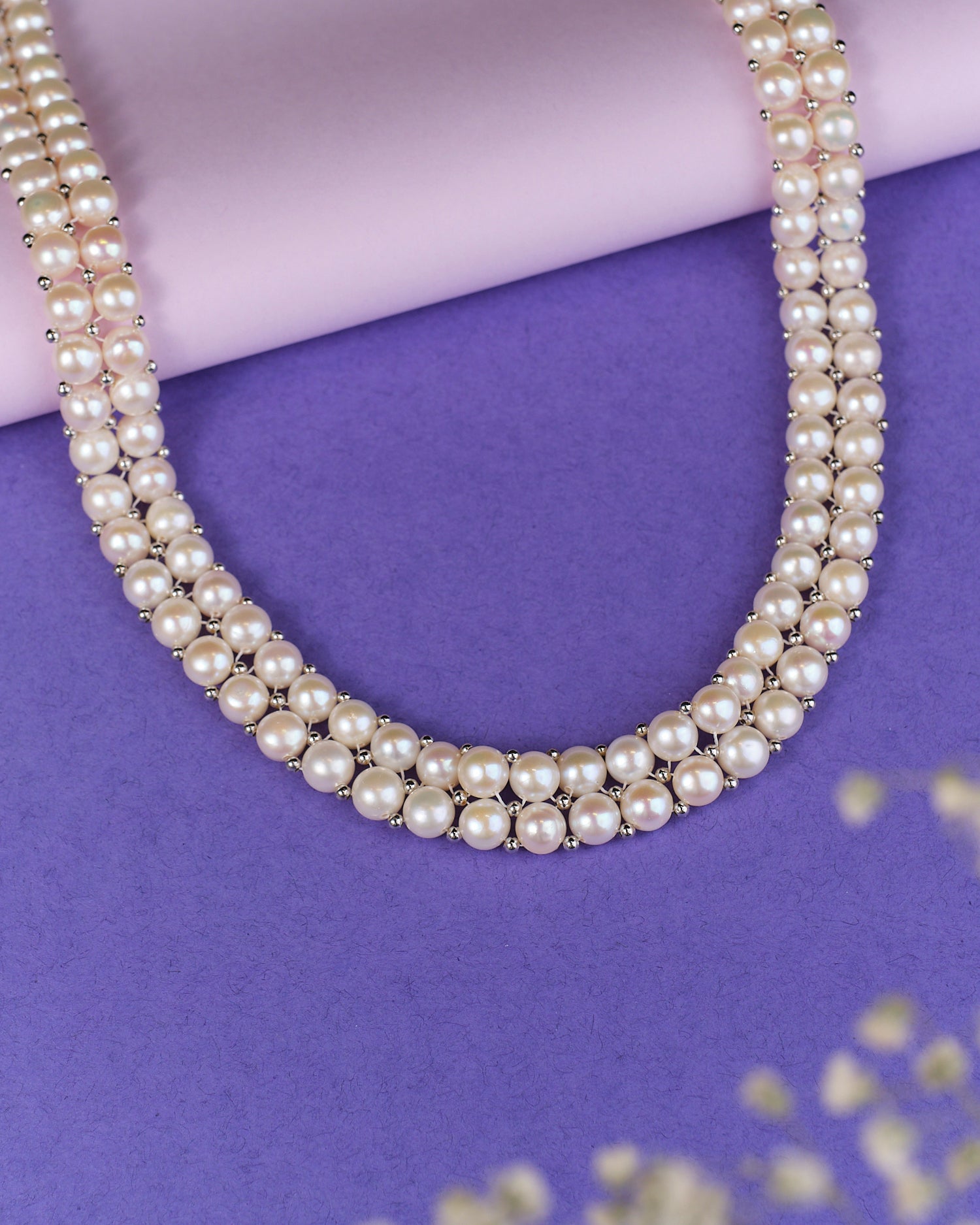 The Wit White Pearl Necklace by Chandrani Pearls elegantly shines against a purple and pink backdrop, accented by small decorative plants.