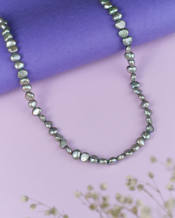 The Rangavati-Full of Colours Pearl Necklace by Chandrani Pearls, featuring stylish irregular gray pearls, is showcased on a purple surface with dried flowers in the foreground.