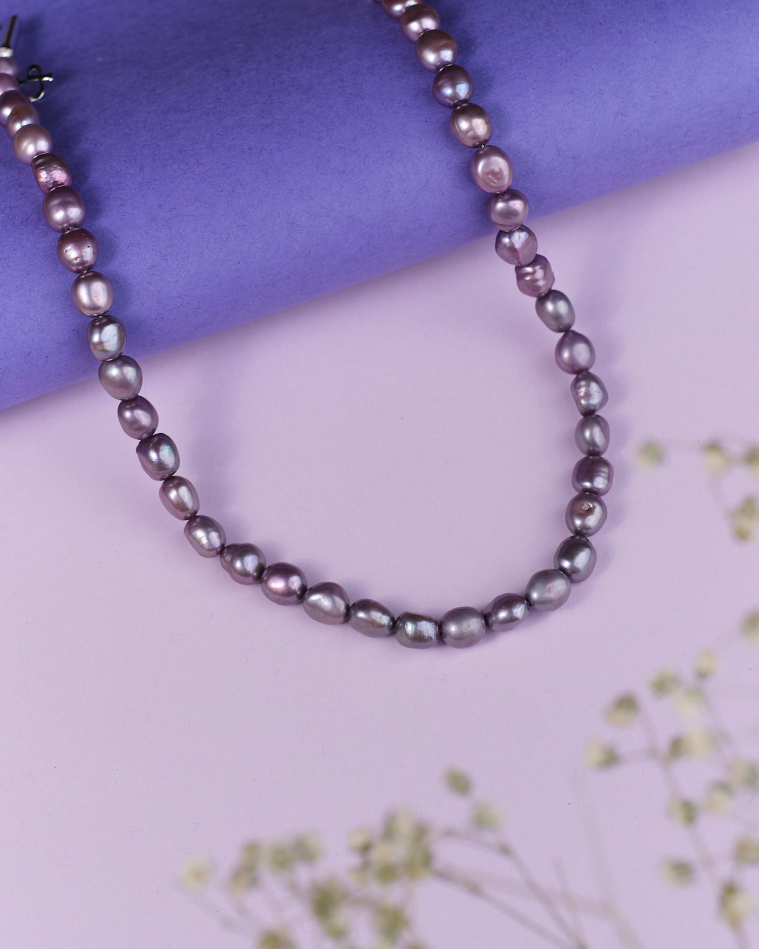 The Splashy Sleet Pearl Necklace by Chandrani Pearls, a blend of purple and pink pearls, elegantly rests on a lavender surface with a blurred floral decoration in the foreground.