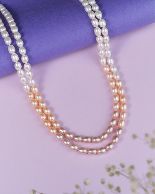 The Bhadrashochi Pearl Necklace by Chandrani Pearls features two luxurious strands, one white and one peach, elegantly arranged on a purple roll atop a pink surface adorned with small corner flowers.