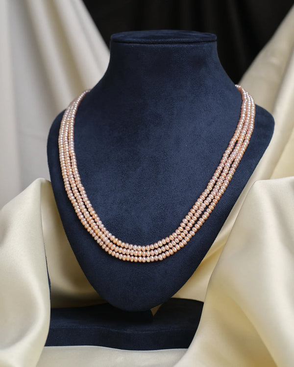 The Chandrani Pearls Freshwater Pink Pearl Necklace, featuring three lustrous strands, is elegantly displayed on a black velvet bust with cream satin fabric accents.