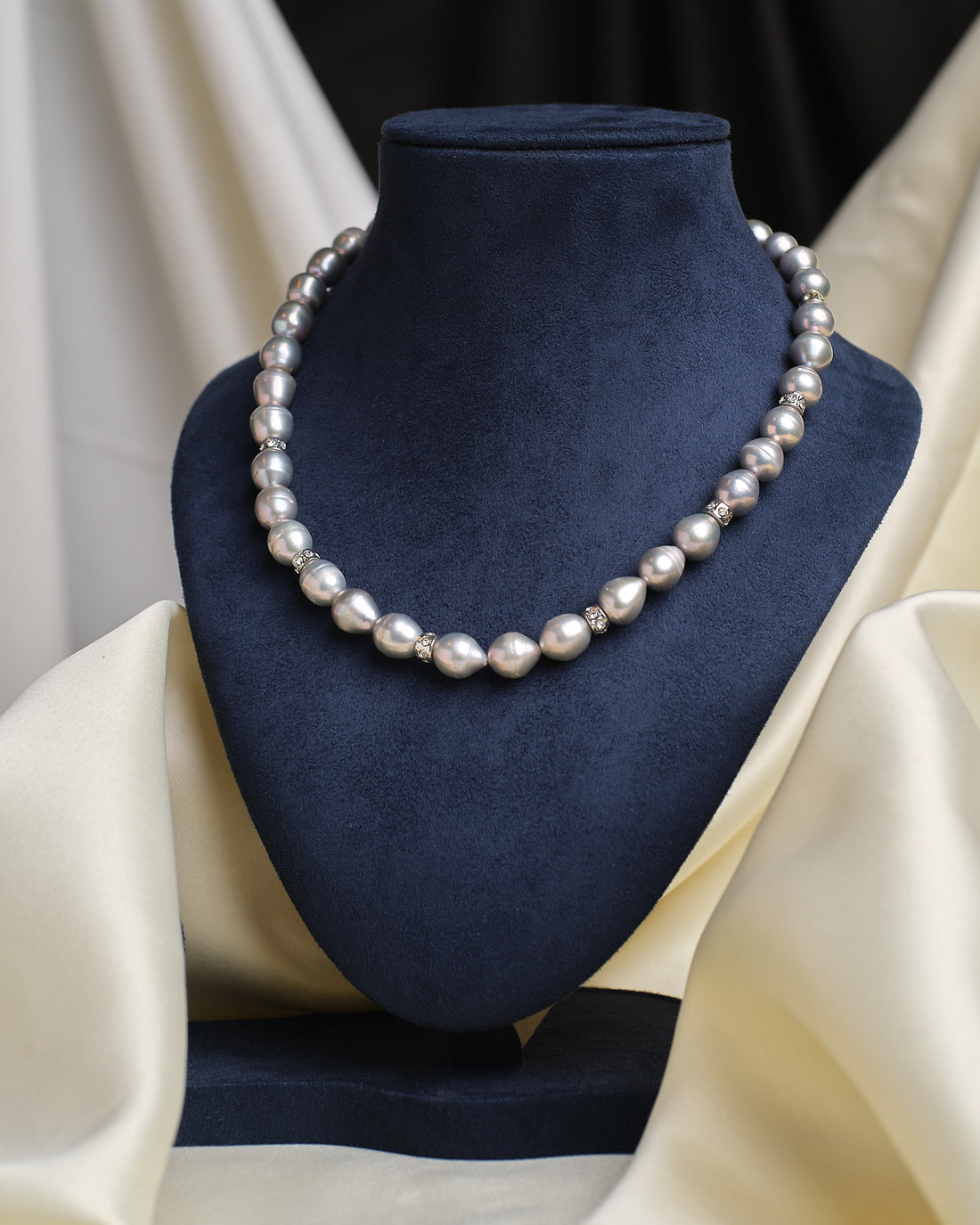 The Baroque Grey Pearl Necklace by Chandrani Pearls features round silver pearls with decorative spacers, highlighting its organic beauty on a dark blue velvet bust draped in cream fabric.