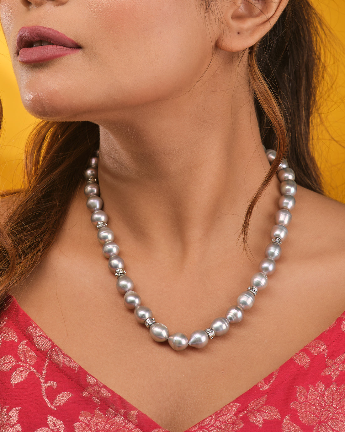 The Baroque Grey Pearl Necklace by Chandrani Pearls features round silver pearls with decorative spacers, highlighting its organic beauty on a dark blue velvet bust draped in cream fabric.