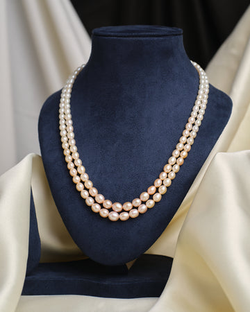 The Graduated Pearl Necklace by Chandrani Pearls, a classic double-stranded design, is displayed on a dark blue velvet bust with cream fabric draped in the background.