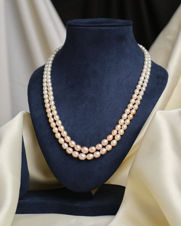 The Graduated Pearl Necklace by Chandrani Pearls, a classic double-stranded design, is displayed on a dark blue velvet bust with cream fabric draped in the background.