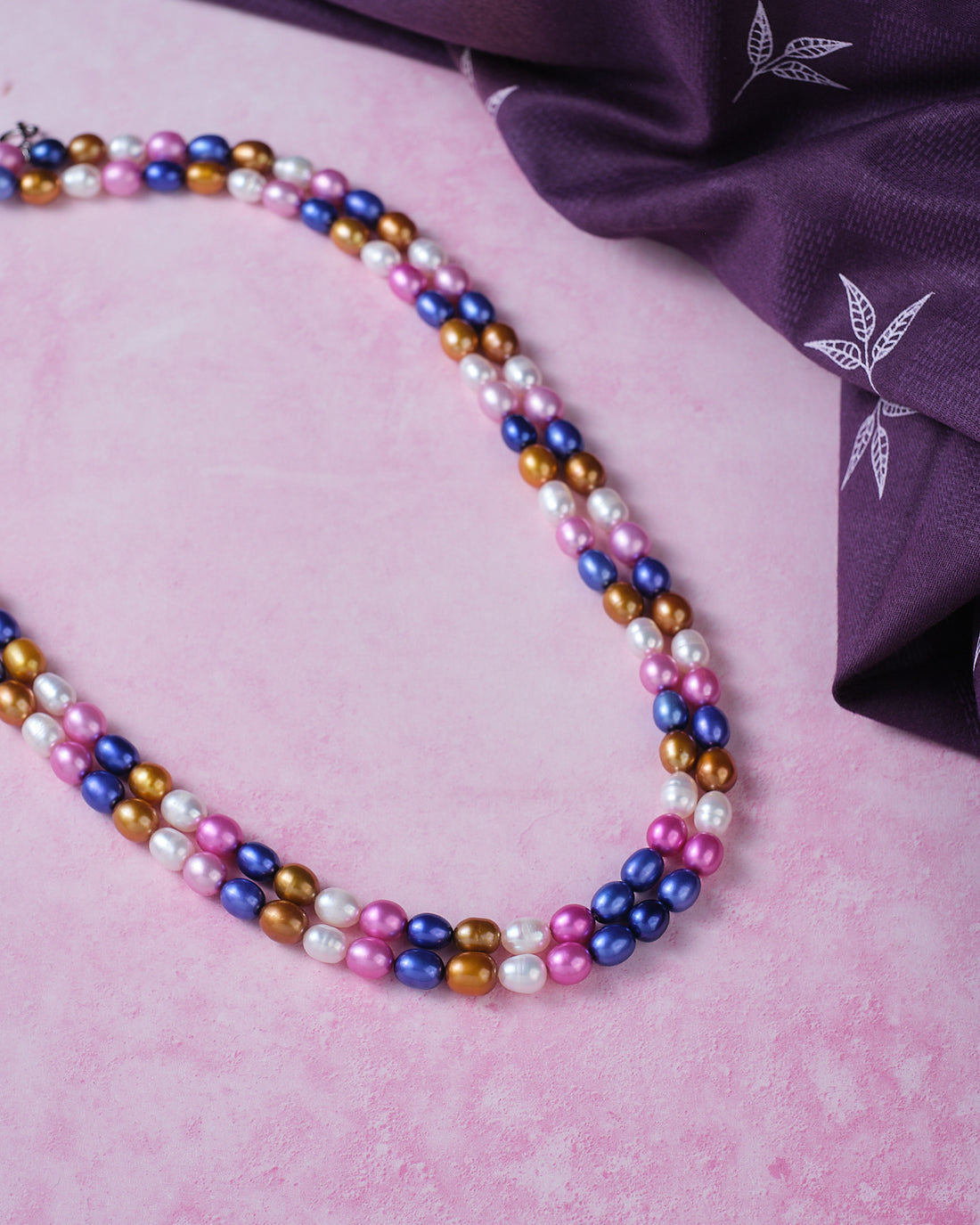 The Multi Colour Pearl Necklace by Chandrani Pearls features blue, pink, purple, and gold pearls arranged on a pink surface beside a folded purple cloth with white leaf patterns.