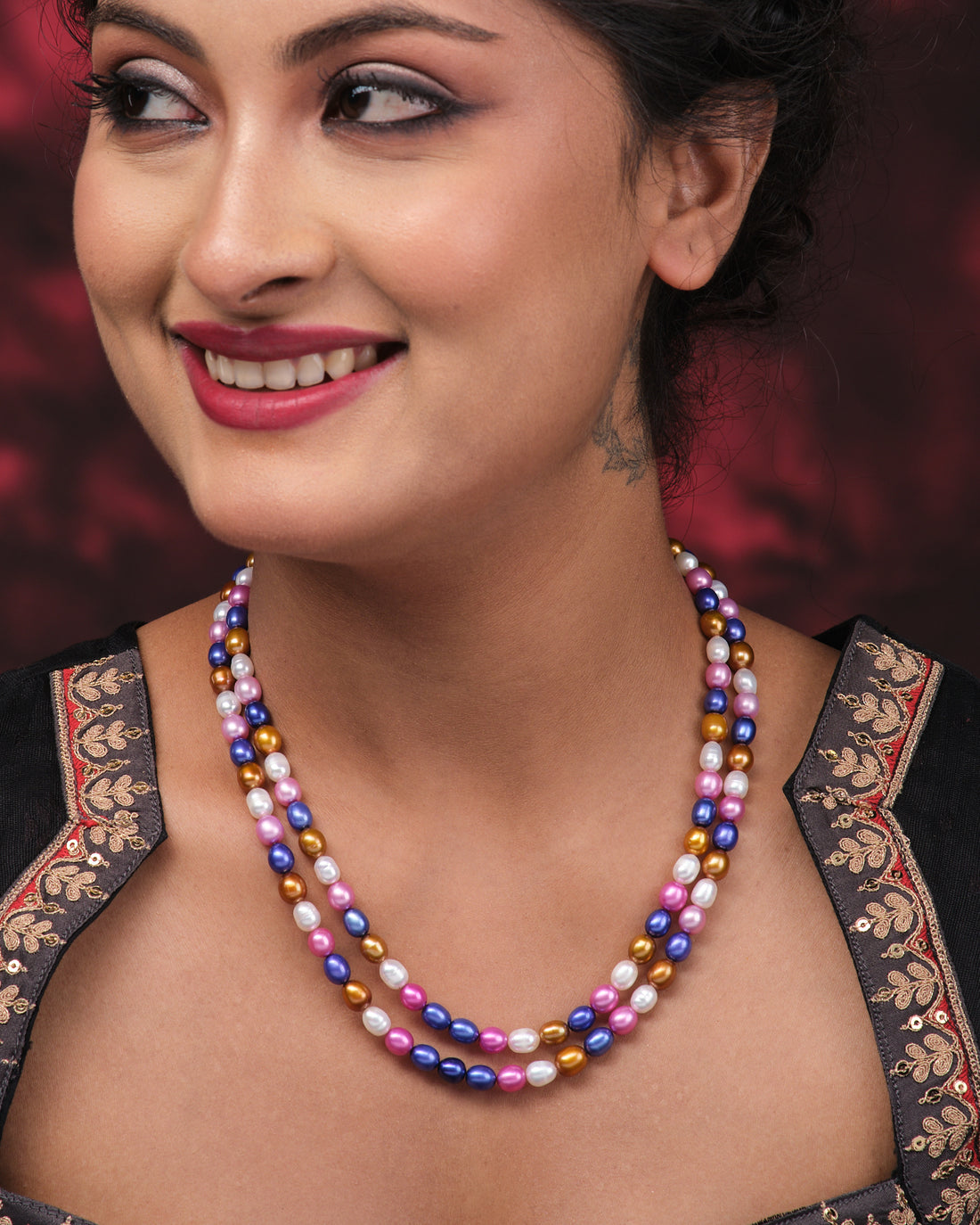 The Multi Colour Pearl Necklace by Chandrani Pearls features blue, pink, purple, and gold pearls arranged on a pink surface beside a folded purple cloth with white leaf patterns.