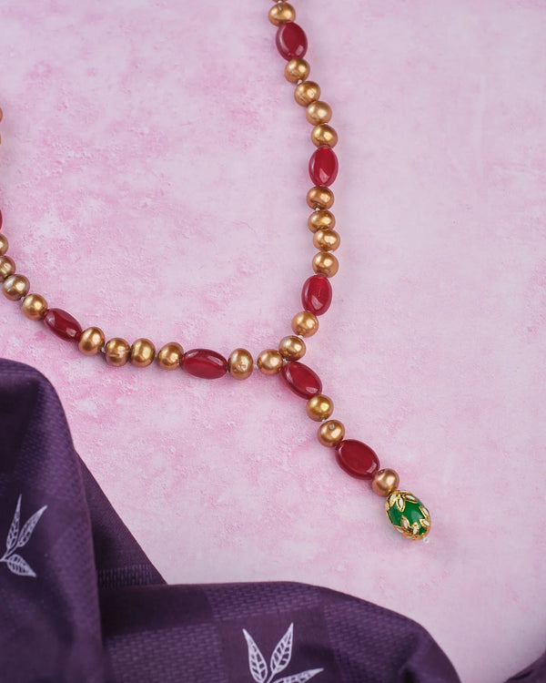 The Elegant Beads & Pearl Mala by Chandrani Pearls—featuring red, gold, and green beads with iridescent pearls—is displayed on a pink surface beside purple fabric adorned with white leaf motifs.