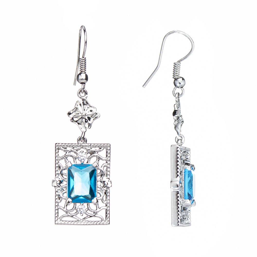Woman with closed eyes and hand on forehead wears the Ocean of Love Earrings by Chandrani Pearls—shimmering blue stones inspired by the ocean—paired with a white blazer against a plain background.
