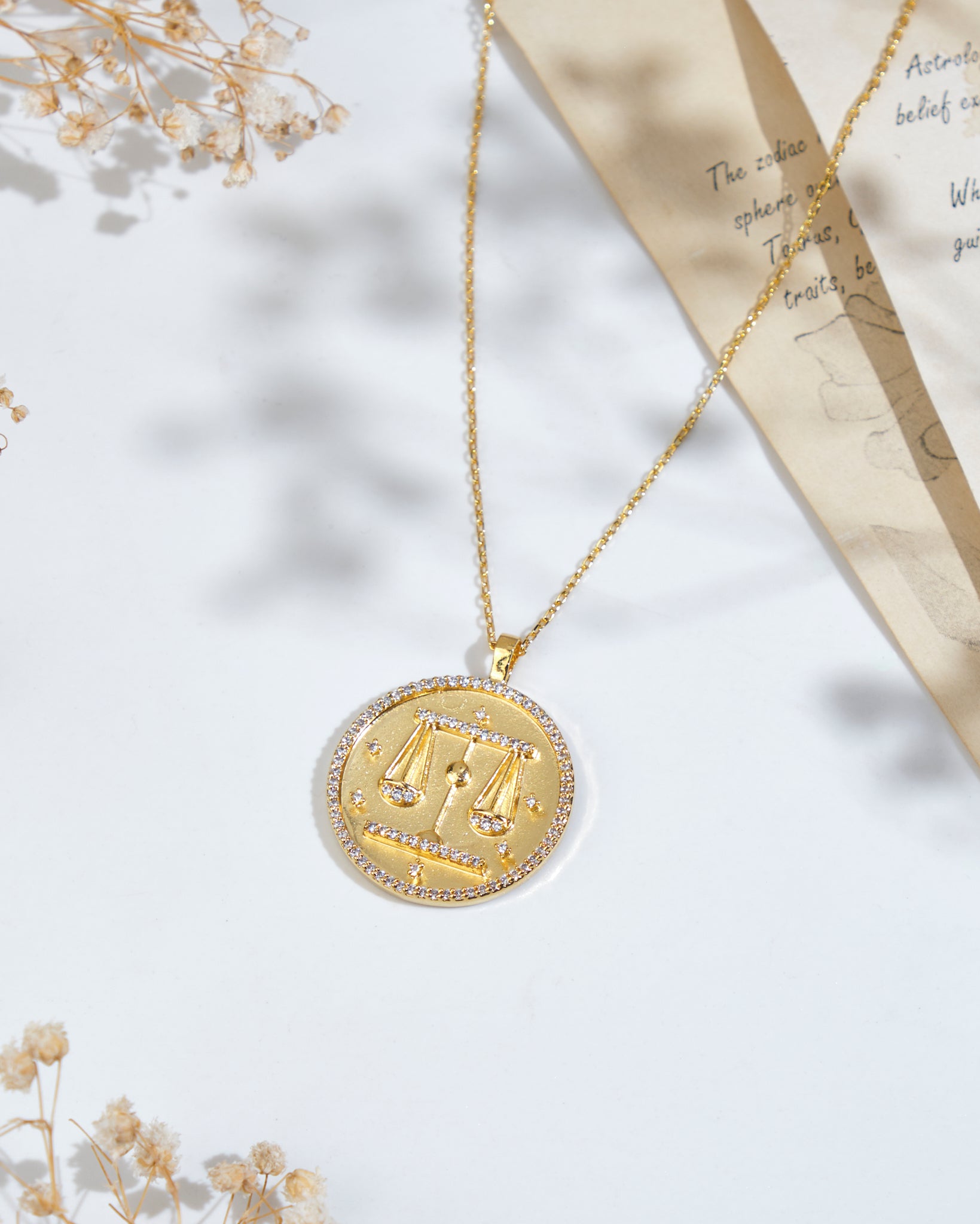 A Soul Of The Scales Libra Pendant by Chandrani Pearls, featuring a scales of justice design, placed on a white surface next to dried flowers and a piece of paper with text and sketches.