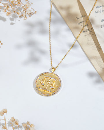 A Soul Of The Scales Libra Pendant by Chandrani Pearls, featuring a scales of justice design, placed on a white surface next to dried flowers and a piece of paper with text and sketches.