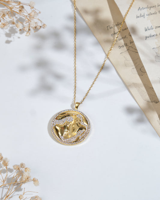 The Soul Of The Twins Gemini Pendant from Chandrani Pearls, a gold necklace featuring a round pendant with an image of a Gemini symbol, accented with small crystals, placed on a white surface alongside a piece of paper and dried flowers.