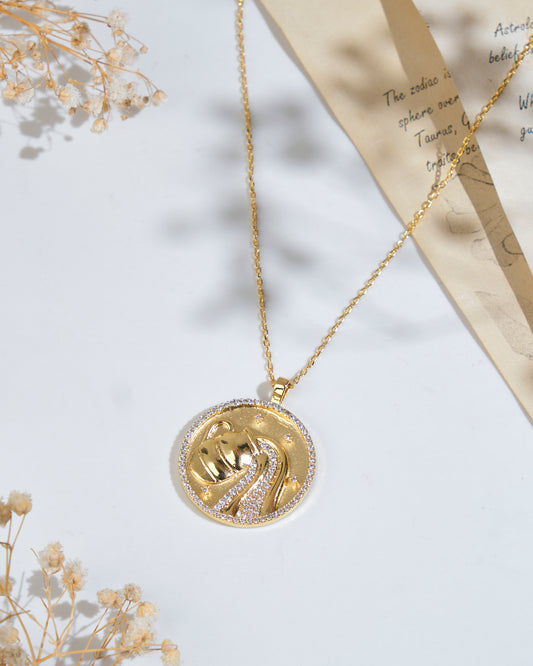 The Soul Of The Water Bearer Aquarius Pendant by Chandrani Pearls, featuring a gold circular design and hanging delicately from a fine chain, is displayed on a white surface next to dried flowers and a handwritten letter.