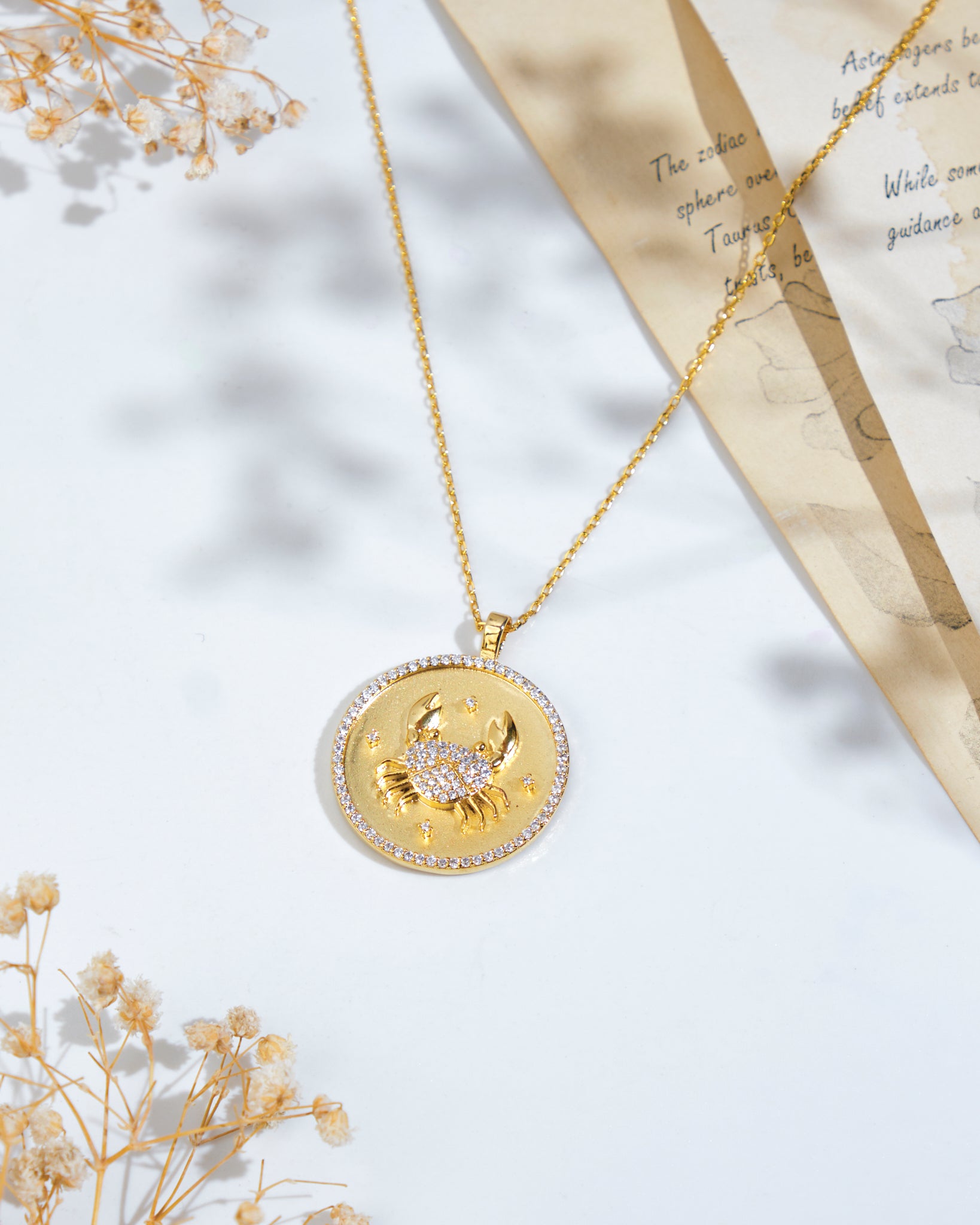 The Soul Of The Crab Cancer Pendant by Chandrani Pearls, a gold necklace featuring a crab emblem, rests on a white surface alongside dried flowers and a partially visible paper with text.