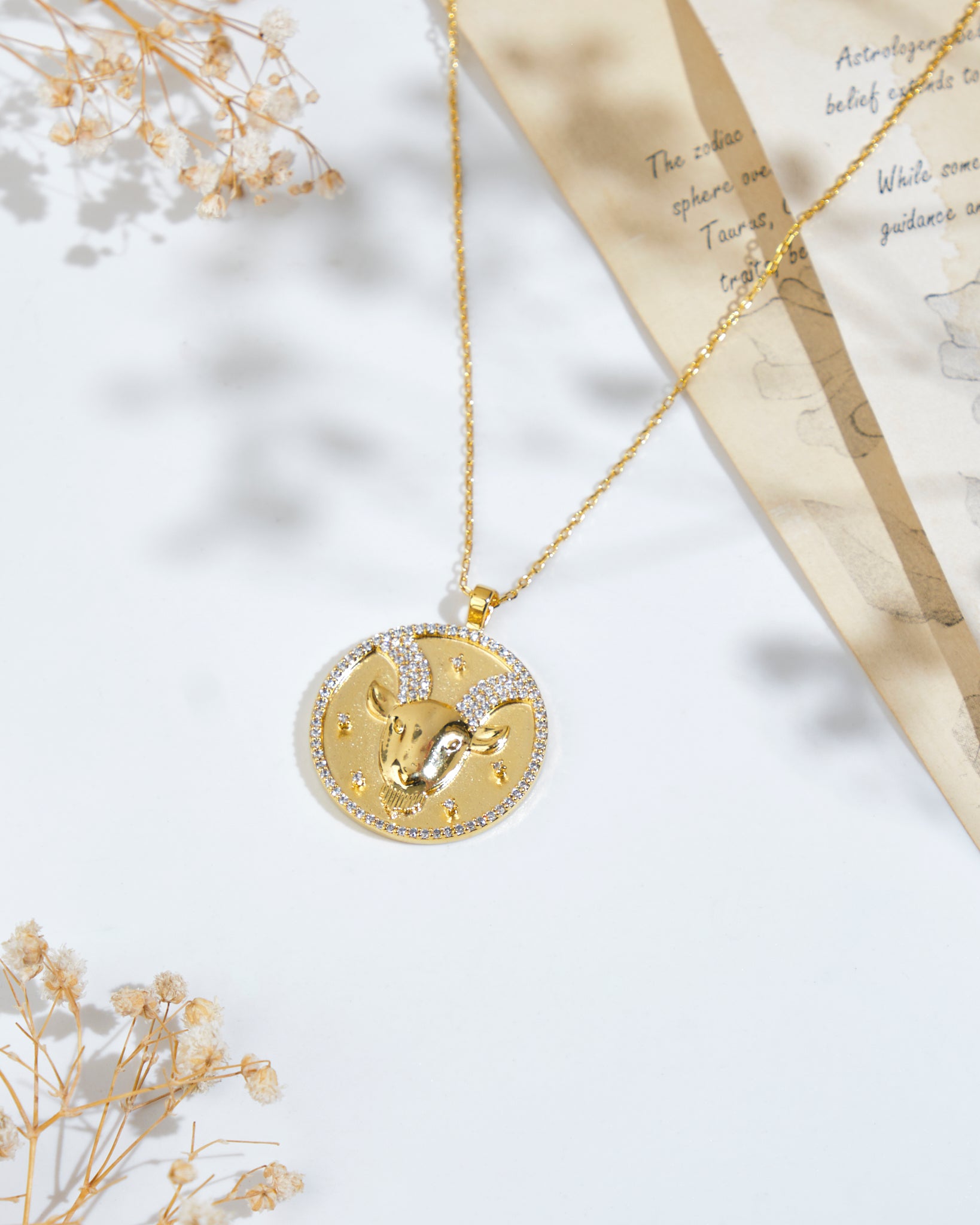 The "Soul Of The Sea Goat Capricorn Pendant" by Chandrani Pearls, a gold necklace with a circular pendant featuring a ram's head, surrounded by small stones, is placed on a white surface with dried flowers and a partially visible zodiac-themed paper.