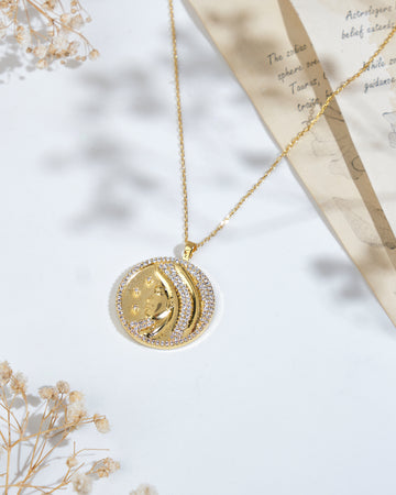 A gold necklace featuring the Soul Of The Virgin Virgo Pendant by Chandrani Pearls, adorned with a moon and star design and surrounded by small crystals, is displayed on a white surface with decorative dried flowers and a partially visible handwritten note.