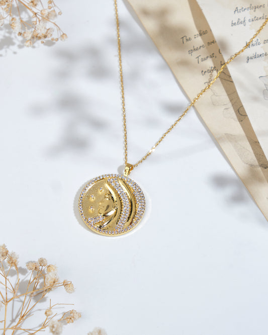 A gold necklace featuring the Soul Of The Virgin Virgo Pendant by Chandrani Pearls, adorned with a moon and star design and surrounded by small crystals, is displayed on a white surface with decorative dried flowers and a partially visible handwritten note.