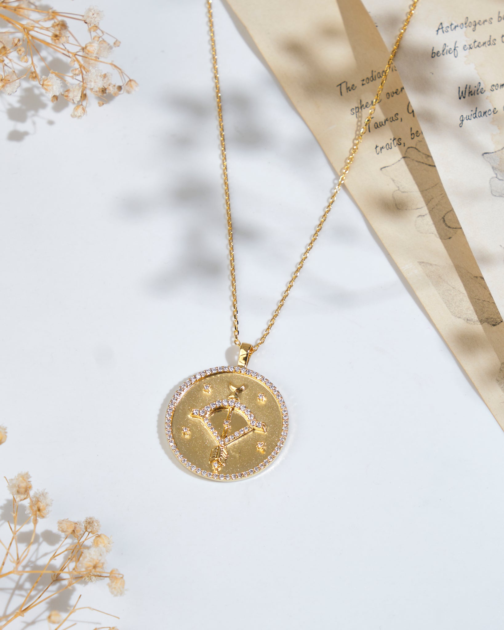 The Soul Of The Archer Sagittarius Pendant by Chandrani Pearls, elegantly displayed on a gold chain, rests on a white surface next to dried flowers and a partially visible piece of paper with text.