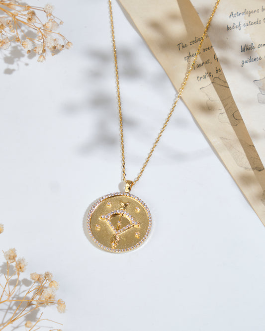 The Soul Of The Archer Sagittarius Pendant by Chandrani Pearls, elegantly displayed on a gold chain, rests on a white surface next to dried flowers and a partially visible piece of paper with text.