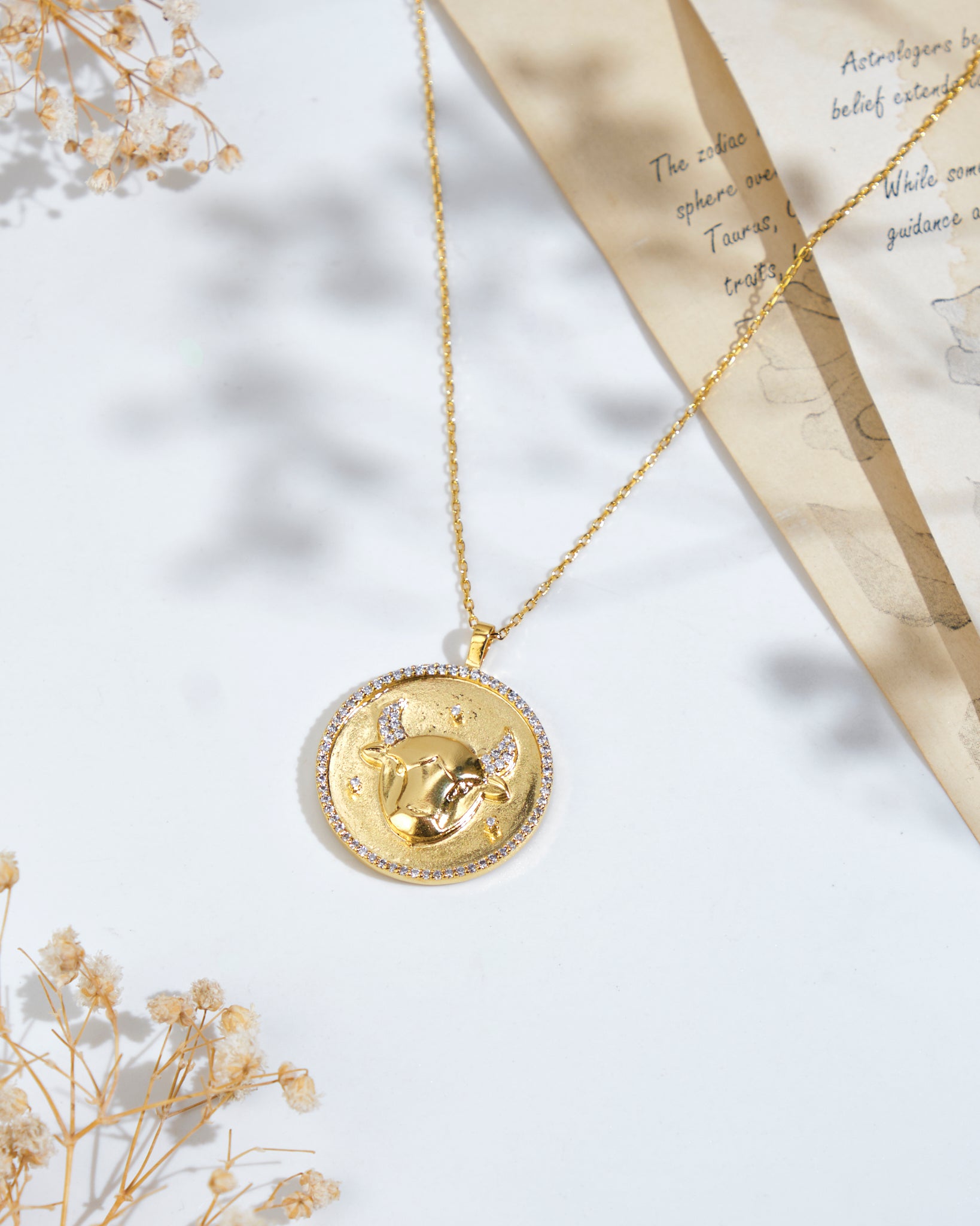 Displayed on a flat surface, the Soul Of The Bull Taurus Pendant from Chandrani Pearls showcases a round pendant with a bull design. Nearby, astrological notes are partially visible on a paper, and dried flowers adorn the scene.