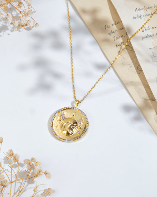 Displayed on a flat surface, the Soul Of The Bull Taurus Pendant from Chandrani Pearls showcases a round pendant with a bull design. Nearby, astrological notes are partially visible on a paper, and dried flowers adorn the scene.