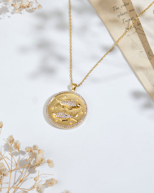 A gold "Soul Of The Fish" Pisces Pendant by Chandrani Pearls, featuring a round design with two fish symbols, lying on a white surface. Dry flowers and a partially visible sheet of paper are in the background.