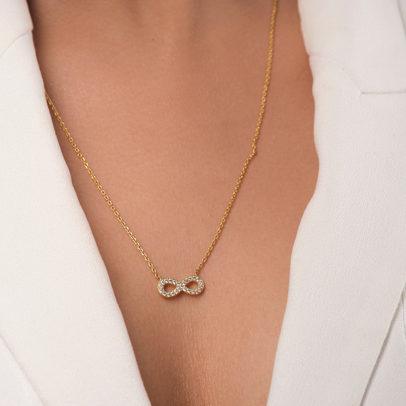 The Undying Love Pendant by Chandrani Pearls, a gold necklace with a diamond-studded infinity symbol, worn over a white blazer, perfectly captures everlasting love and timeless elegance.