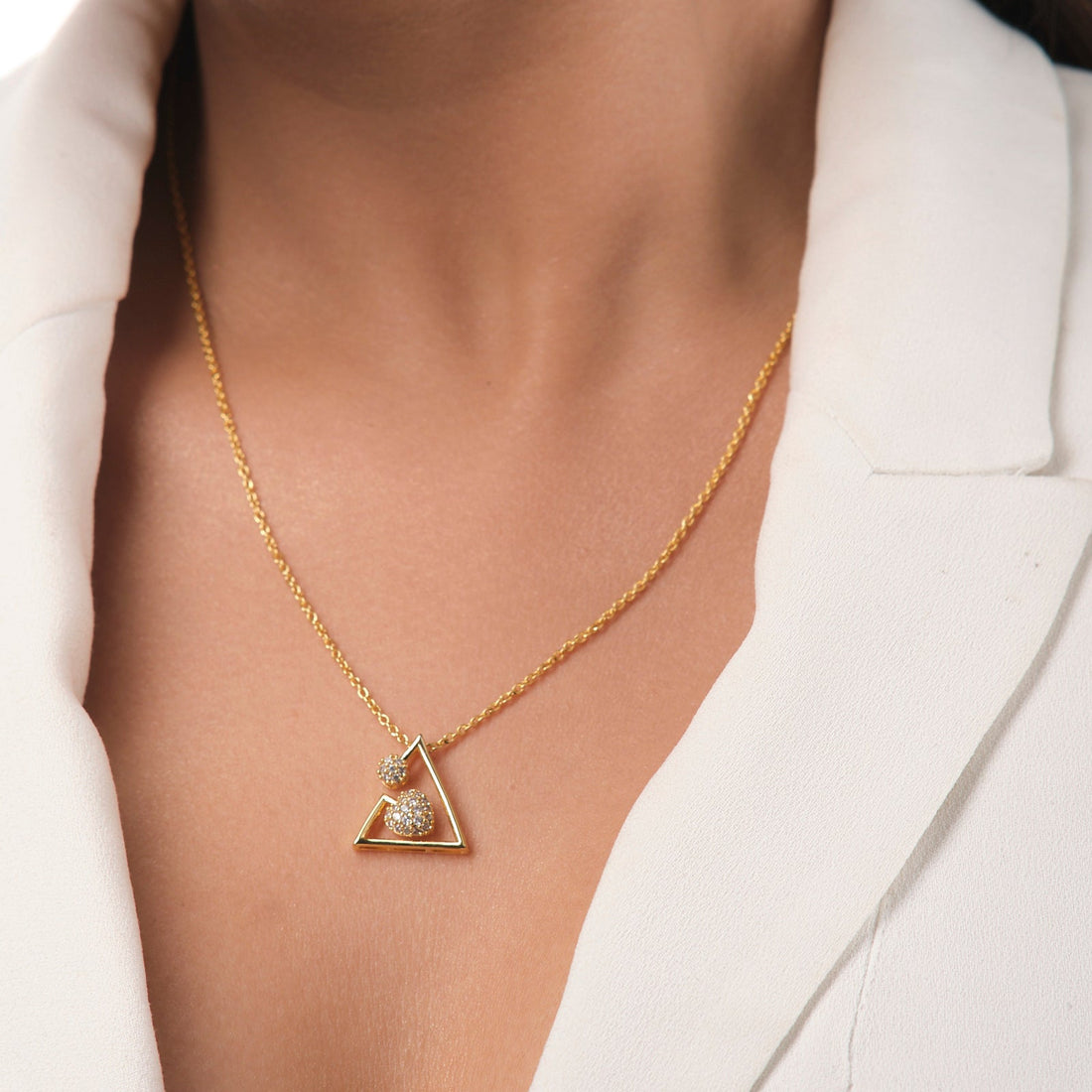 A person in a white blazer wears Chandrani Pearls’ Grateful Heart Pendant—a gold necklace with a triangular motif, symbolizing a mother's love and adorned with small decorative stones.