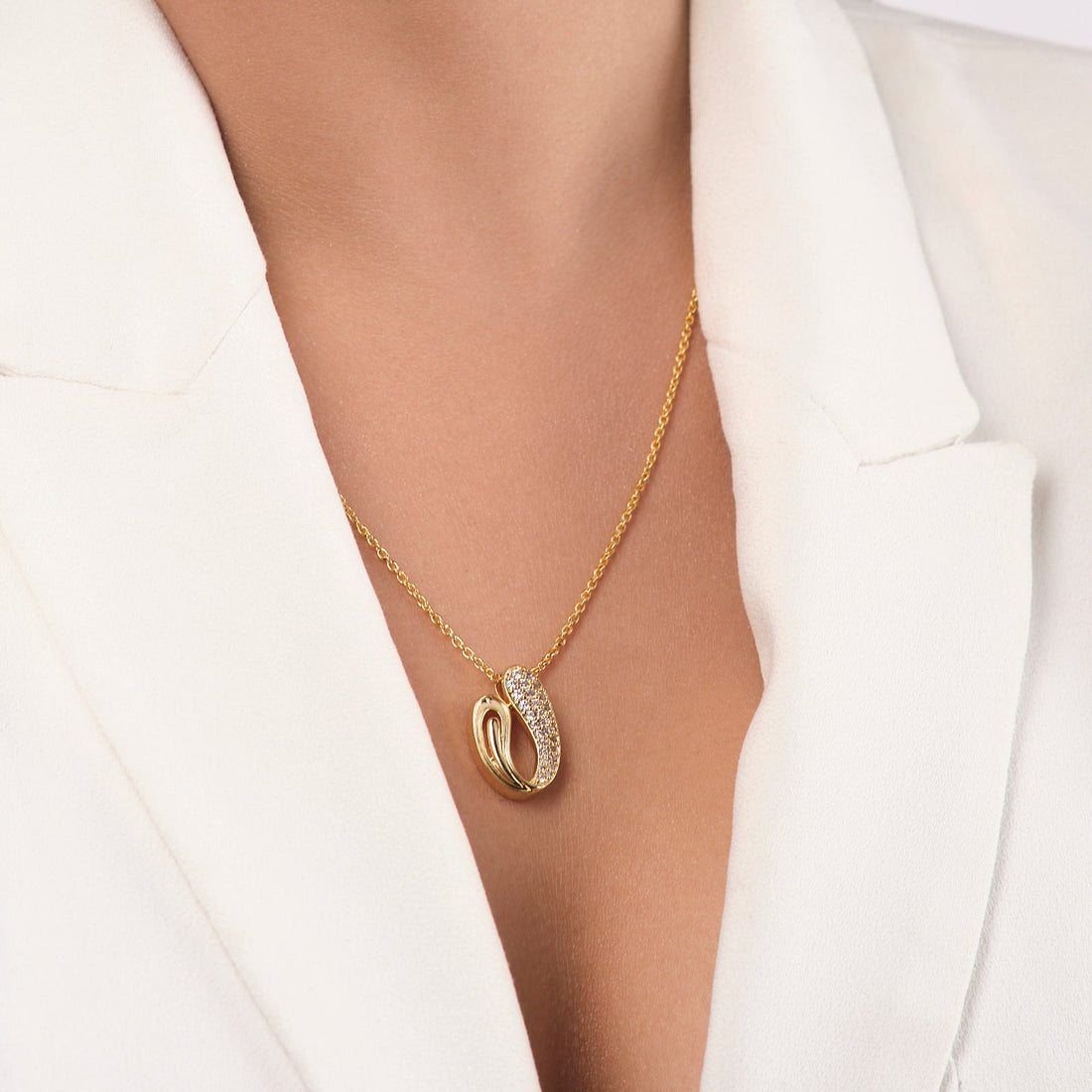 A person wearing a white blazer showcases the Due Drop Glimmer Pendant by Chandrani Pearls—a gold chain necklace with two interlocking rings (one plain, one stone-studded) and a delicate teardrop pearl symbolizing mother's love.