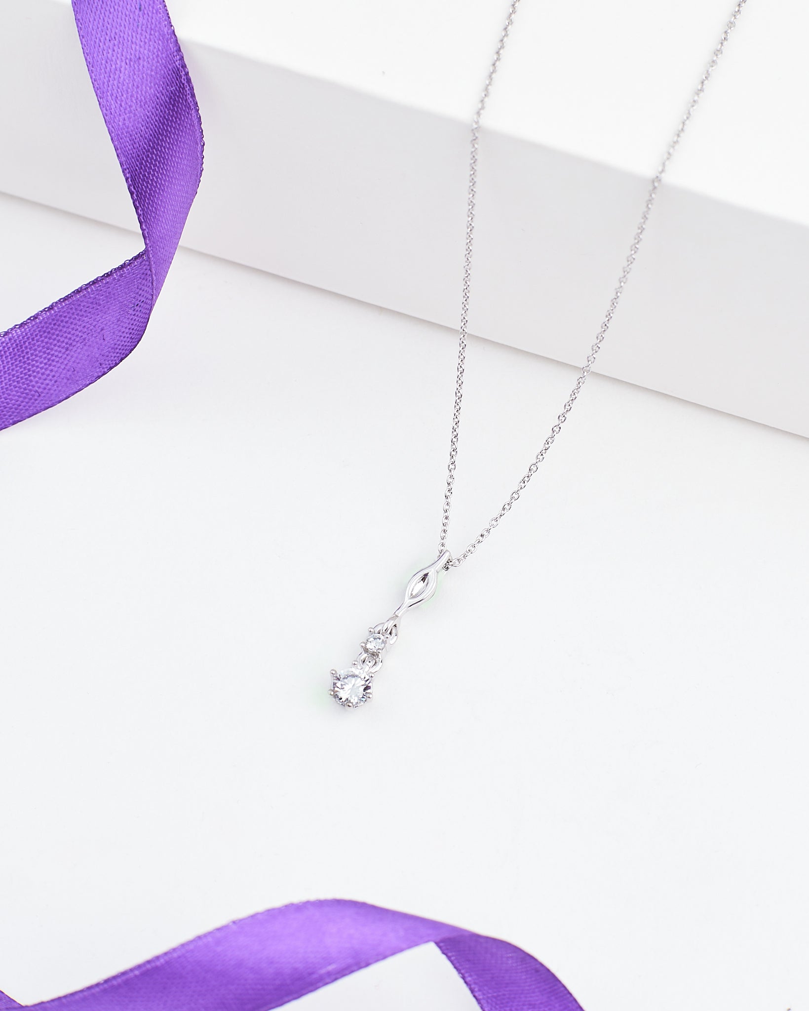 The Elegant CZ Pendant in Demifine Silver by Chandrani Pearls showcases a clear cubic zirconia infinity design, presented on a white surface with a purple ribbon.