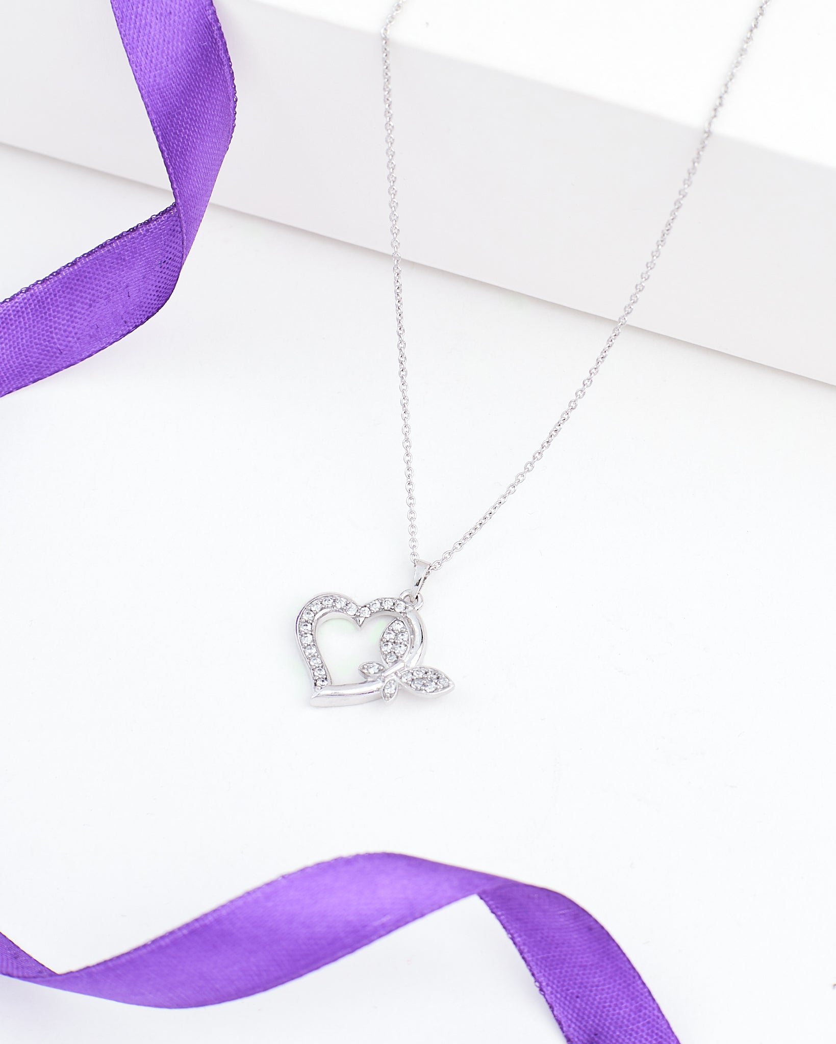 The Chandrani Pearls Heart-Butterfly Demifine Silver CZ Pendant features a heart-butterfly shape with sparkling crystals and leaf detail, shown on a white surface with a purple ribbon.
