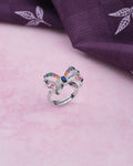 The Whimsical Colorful Butterfly Ring by Chandrani Pearls, featuring playful elegance and multicolored gemstones, is displayed on a pink surface beside folded purple leaf-patterned fabric—a stunning accessory.