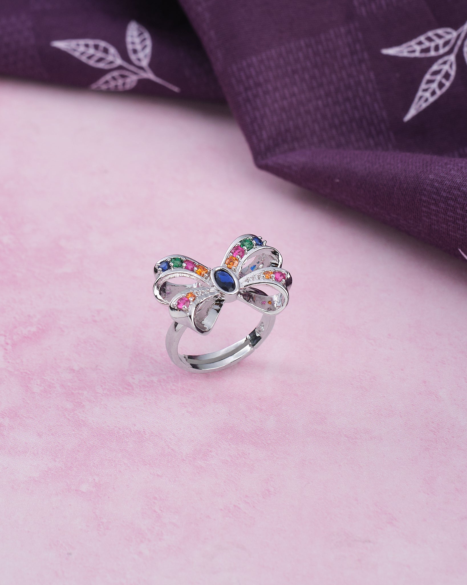 The Whimsical Colorful Butterfly Ring by Chandrani Pearls, featuring playful elegance and multicolored gemstones, is displayed on a pink surface beside folded purple leaf-patterned fabric—a stunning accessory.
