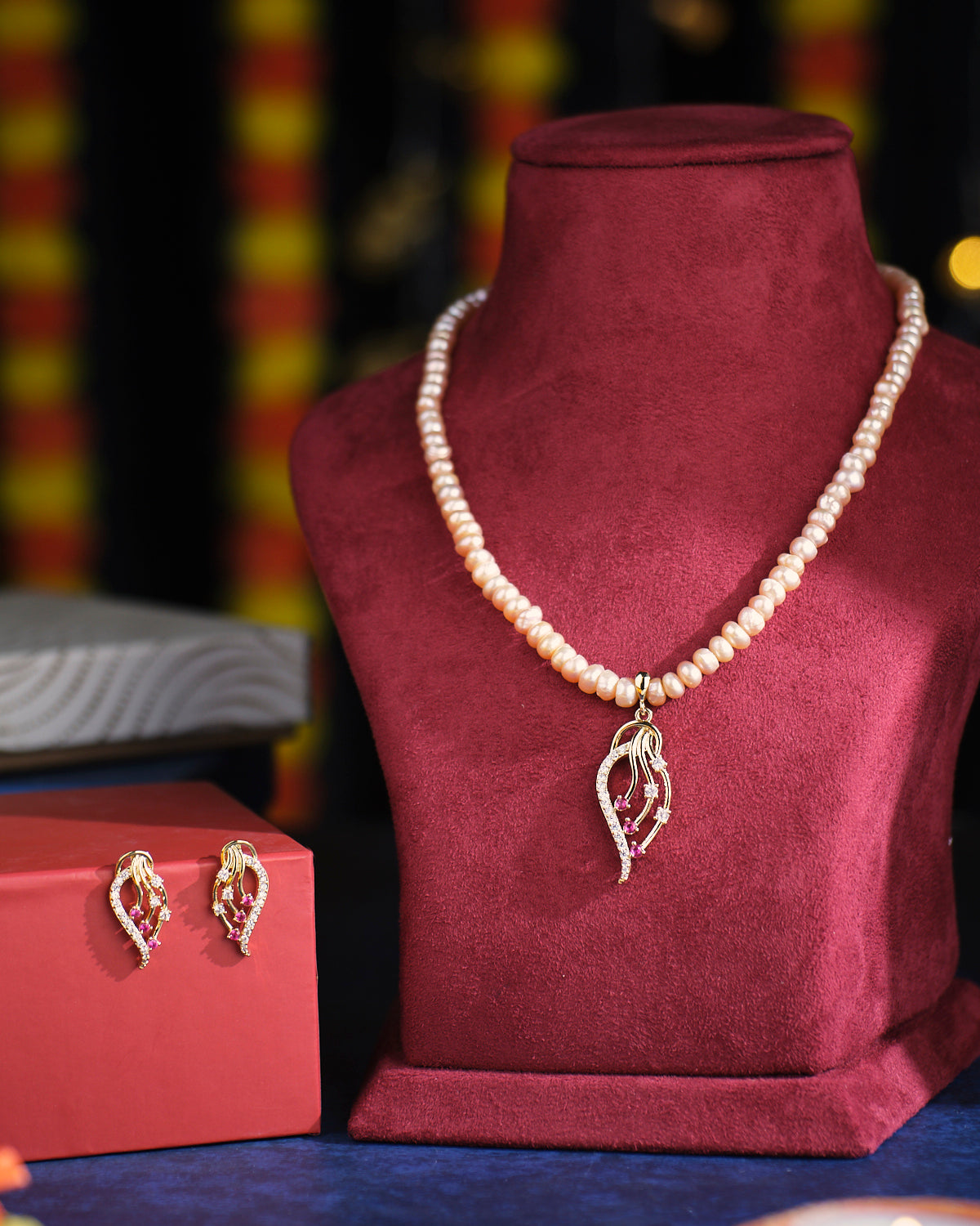 The Windy Leaf Pearl Necklace Set by Chandrani Pearls, featuring a gold and diamond pendant, is elegantly displayed on a red velvet stand. Nearby, matching earrings are showcased atop a red box.