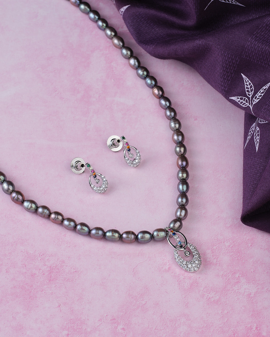 The Chandrani Pearls Vintage Black Stone Simple Pearl Necklace Set, featuring a crystal pendant and matching stud earrings, is displayed on a pink surface beside a folded purple patterned cloth.