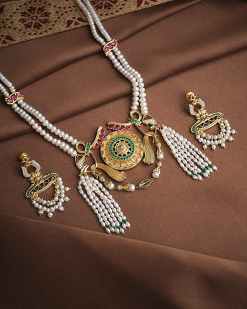 The Heritage Royal Pearl Set by Chandrani Pearls, featuring a gold, green, and pearl necklace with matching earrings, is displayed on a brown fabric background.