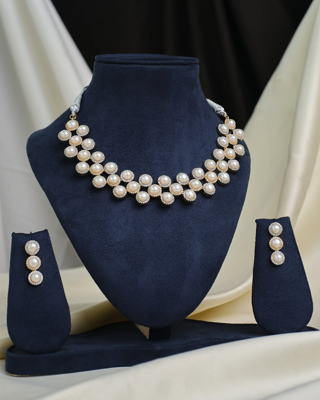 The CZ Infinity Necklace Set by Chandrani Pearls features a pearl necklace, matching pearl earrings, and a CZ-studded infinity pendant, displayed on a navy blue stand with cream and black fabric in the background.