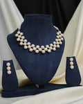 The CZ Infinity Necklace Set by Chandrani Pearls features a pearl necklace, matching pearl earrings, and a CZ-studded infinity pendant, displayed on a navy blue stand with cream and black fabric in the background.
