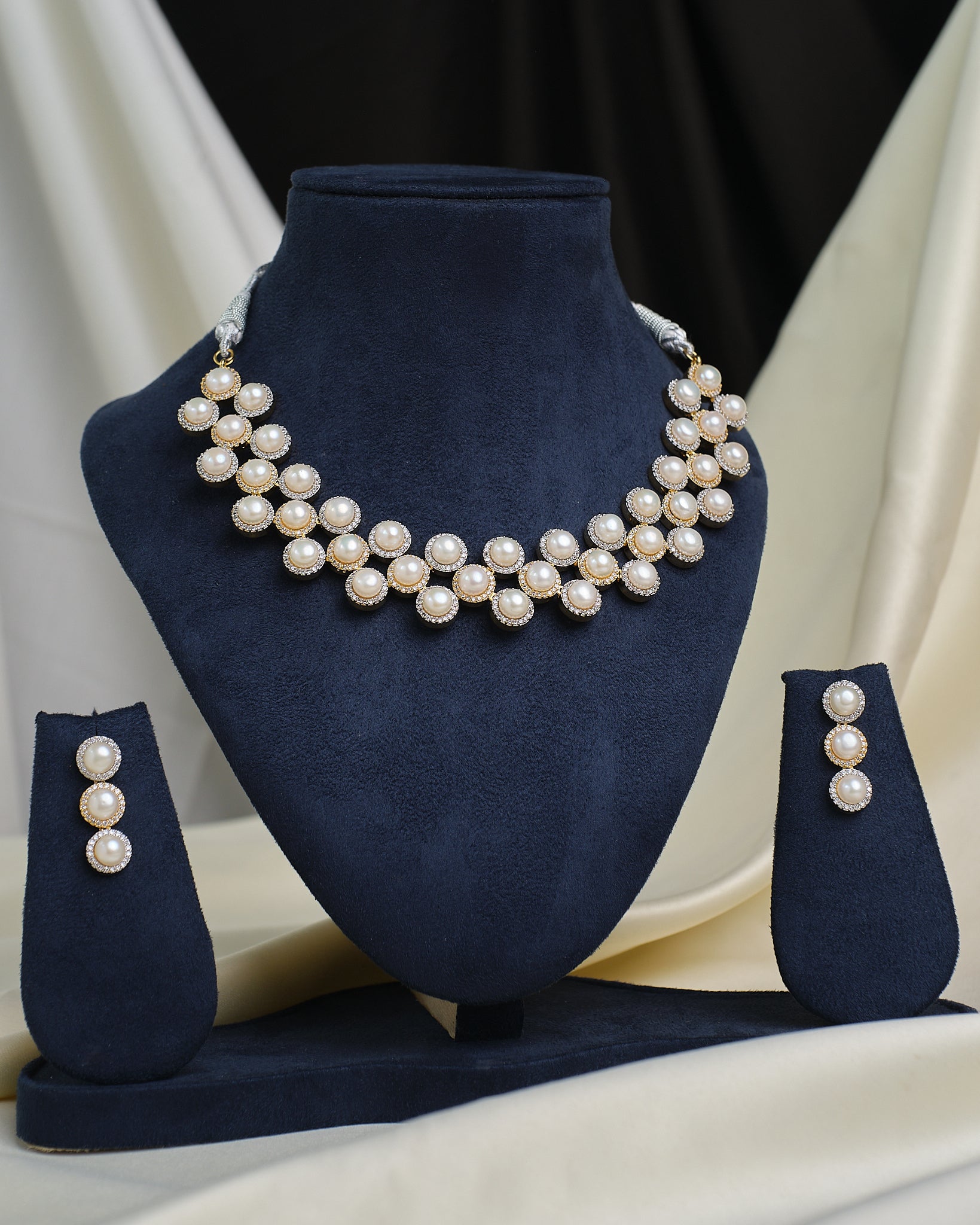 The CZ Infinity Necklace Set by Chandrani Pearls features a pearl necklace, matching pearl earrings, and a CZ-studded infinity pendant, displayed on a navy blue stand with cream and black fabric in the background.