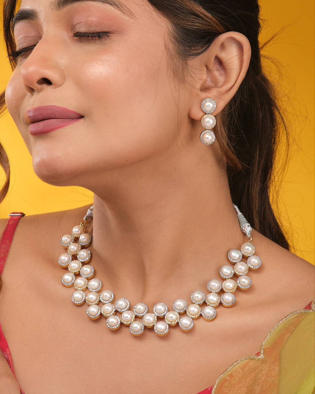 The CZ Infinity Necklace Set by Chandrani Pearls features a pearl necklace, matching pearl earrings, and a CZ-studded infinity pendant, displayed on a navy blue stand with cream and black fabric in the background.