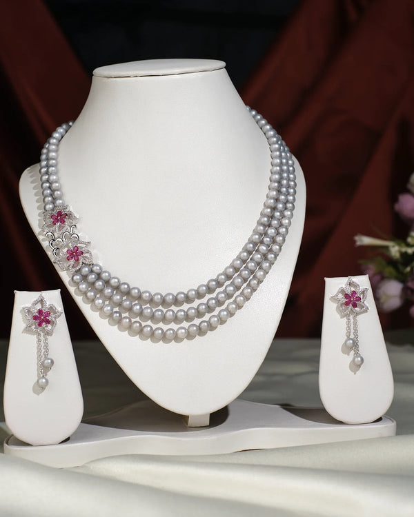The Ravishing Pearl Necklace Set by Chandrani Pearls India features three pearl strands with silver and pink floral accents, displayed with matching pearl and pink stone earrings on a white jewelry stand.
