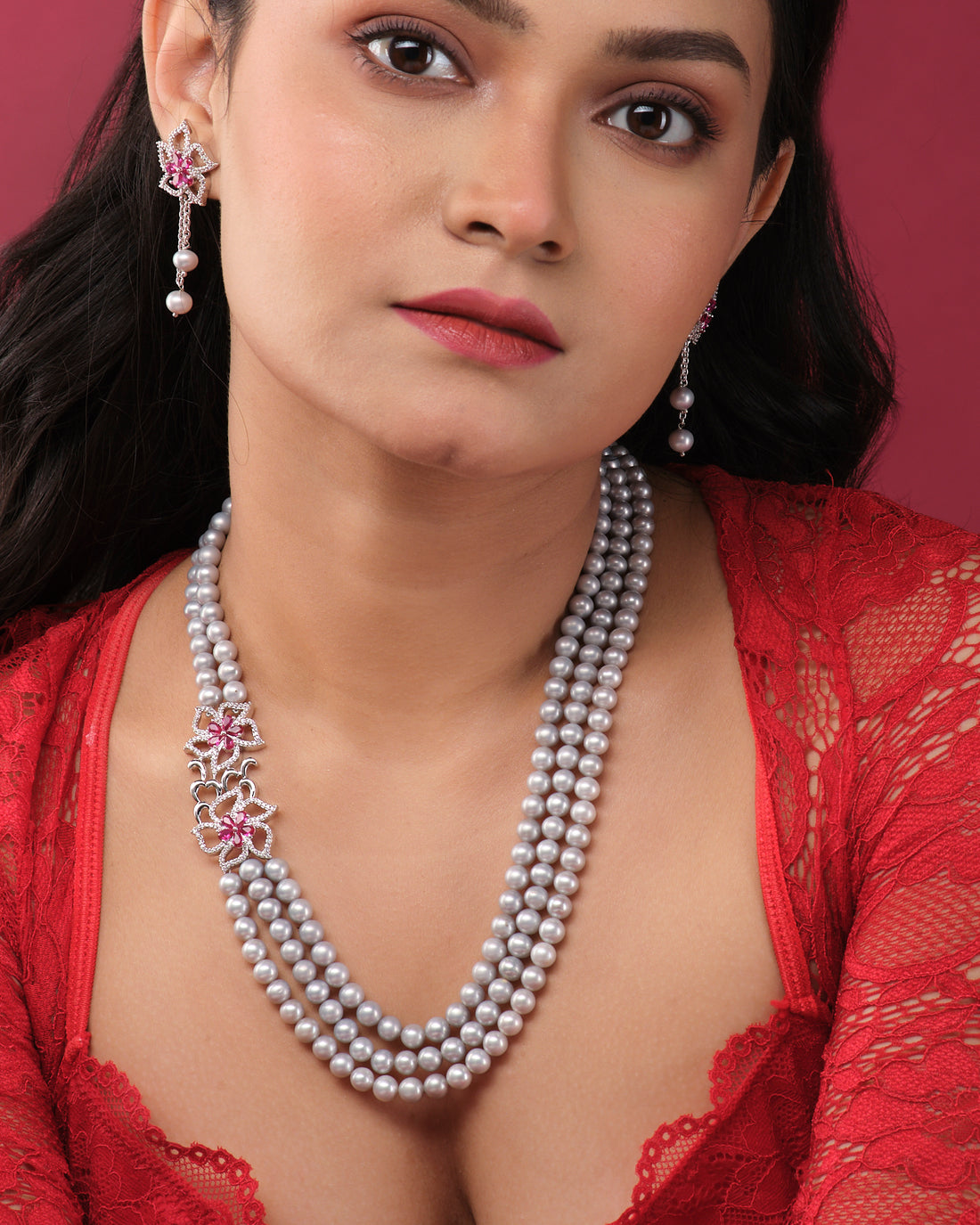 The Ravishing Pearl Necklace Set by Chandrani Pearls India features three pearl strands with silver and pink floral accents, displayed with matching pearl and pink stone earrings on a white jewelry stand.
