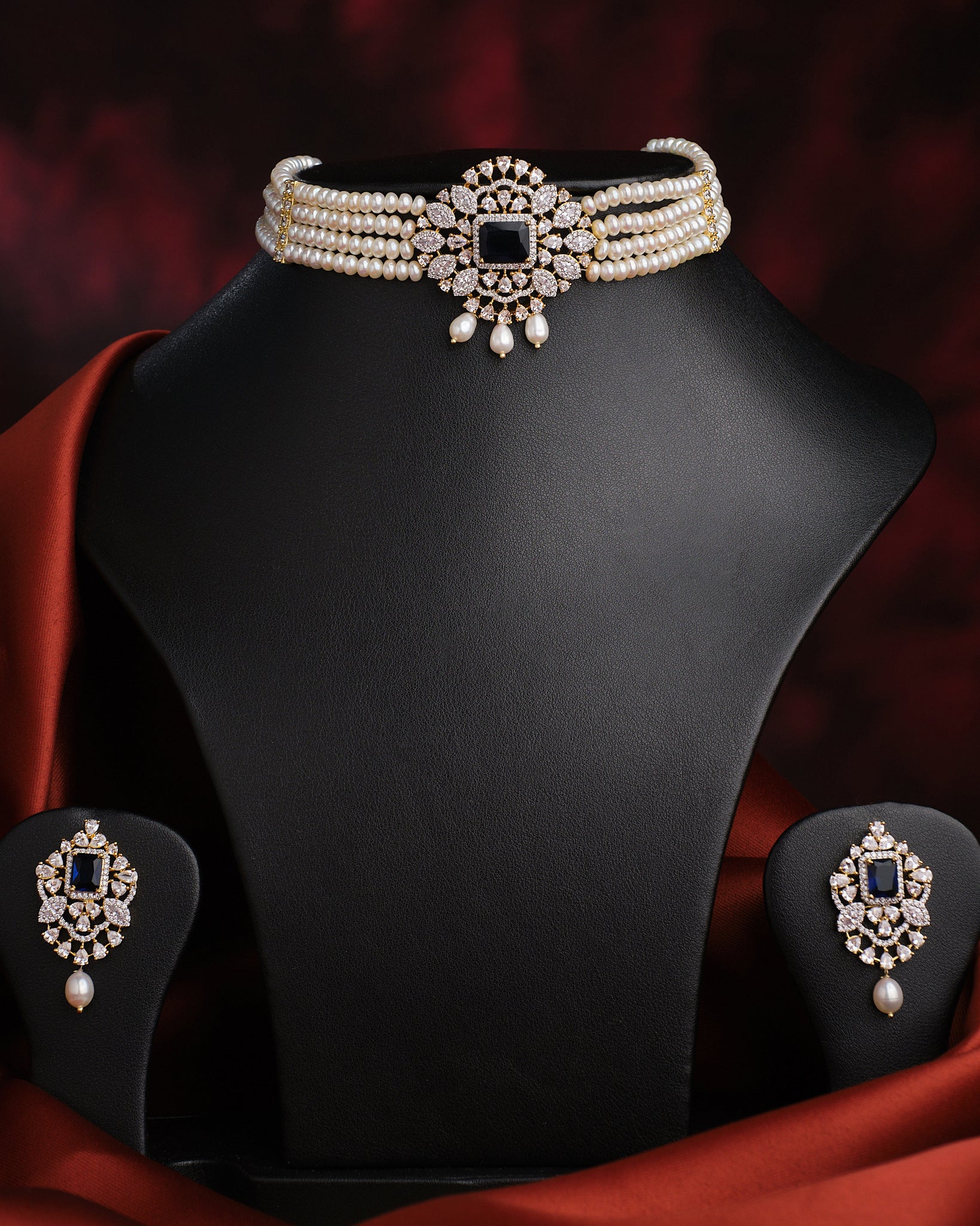 The Newfangled Pearl Necklace Set by Chandrani Pearls, featuring white pearls and gold disc accents with matching earrings, is showcased on a black jewelry stand against a dark red background.