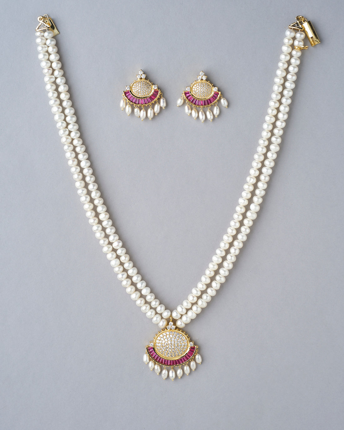 The Traditional Pearl Necklace Set by Chandrani Pearls India features a gold and pink pendant displayed on a white stand, with matching earrings on separate stands—a perfect addition to any jewelry collection.