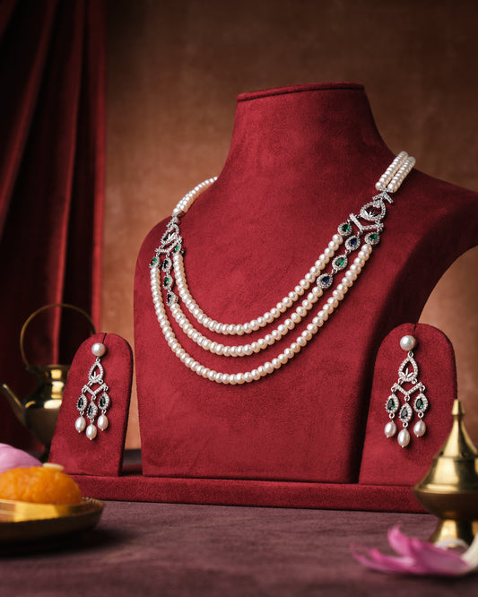 A display of the Swirly Dual Side CZ Pendant Pearl Necklace Set by Chandrani Pearls India showcased on a red velvet stand with a brown velvet background, accompanied by decorative items and floral accents.