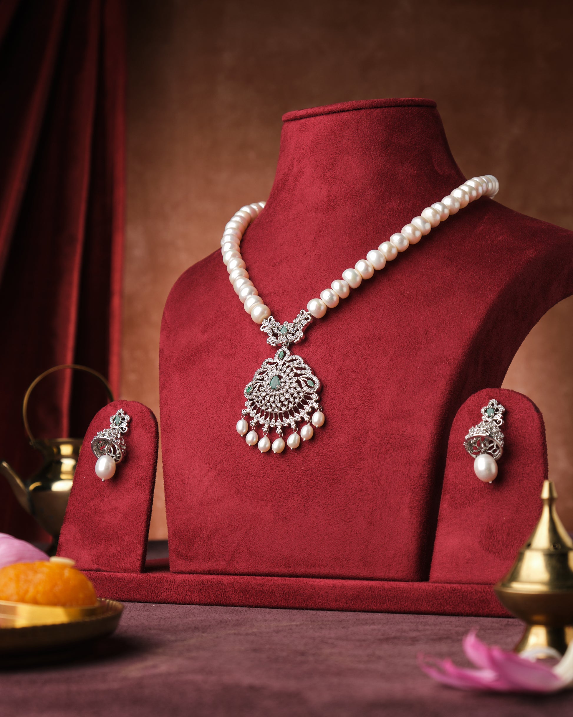 The Meghraa CZ Pendant Pearl Necklace Set by Chandrani Pearls India, featuring matching earrings, is displayed on a red velvet bust against a brown backdrop.