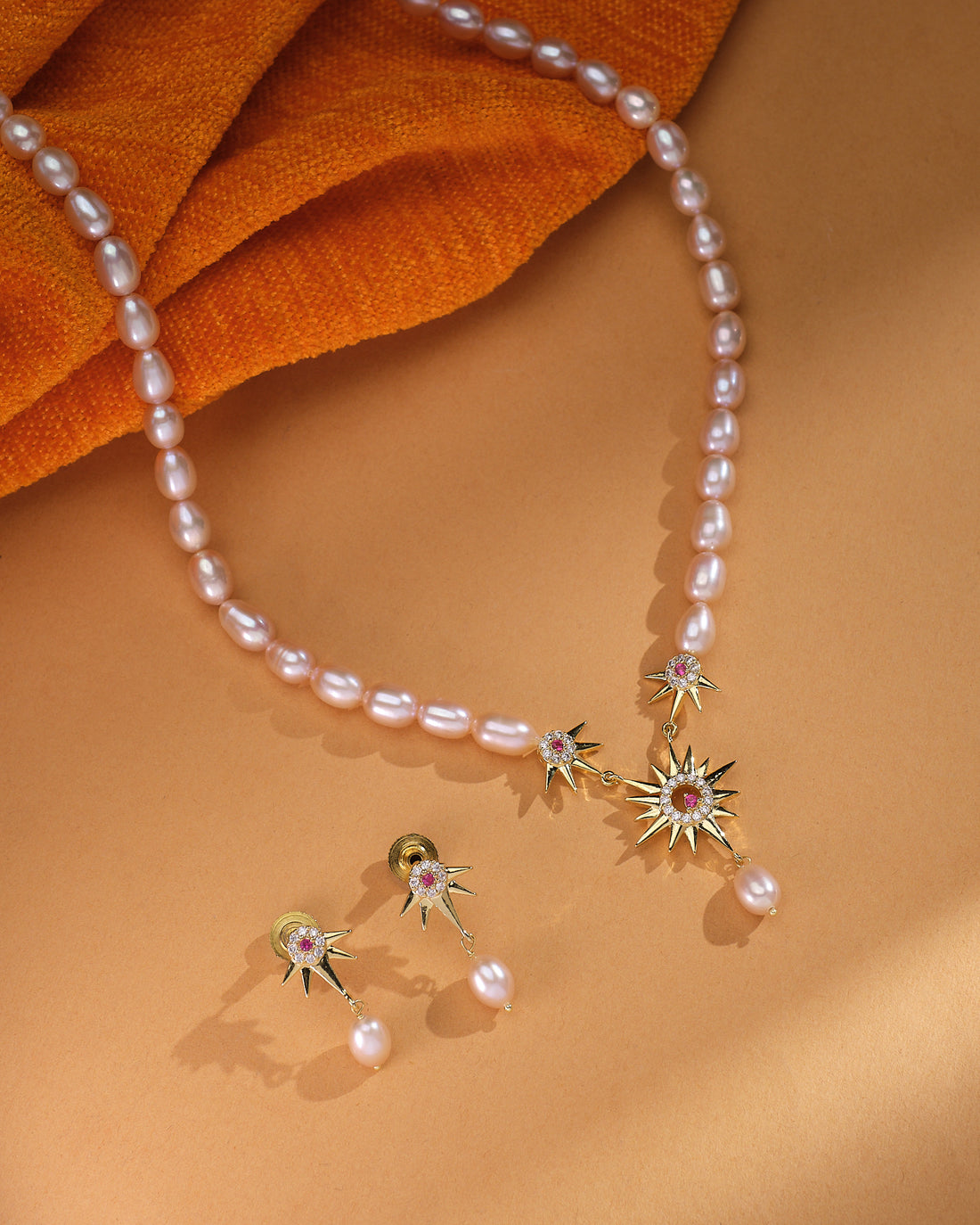 The Zara Gold Layered Set by Chandrani Pearls features a pearl necklace with gold starburst pendants and matching minimalist pearl drop studs, elegantly displayed on a tan surface with orange fabric.