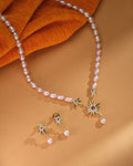 The Zara Gold Layered Set by Chandrani Pearls features a pearl necklace with gold starburst pendants and matching minimalist pearl drop studs, elegantly displayed on a tan surface with orange fabric.