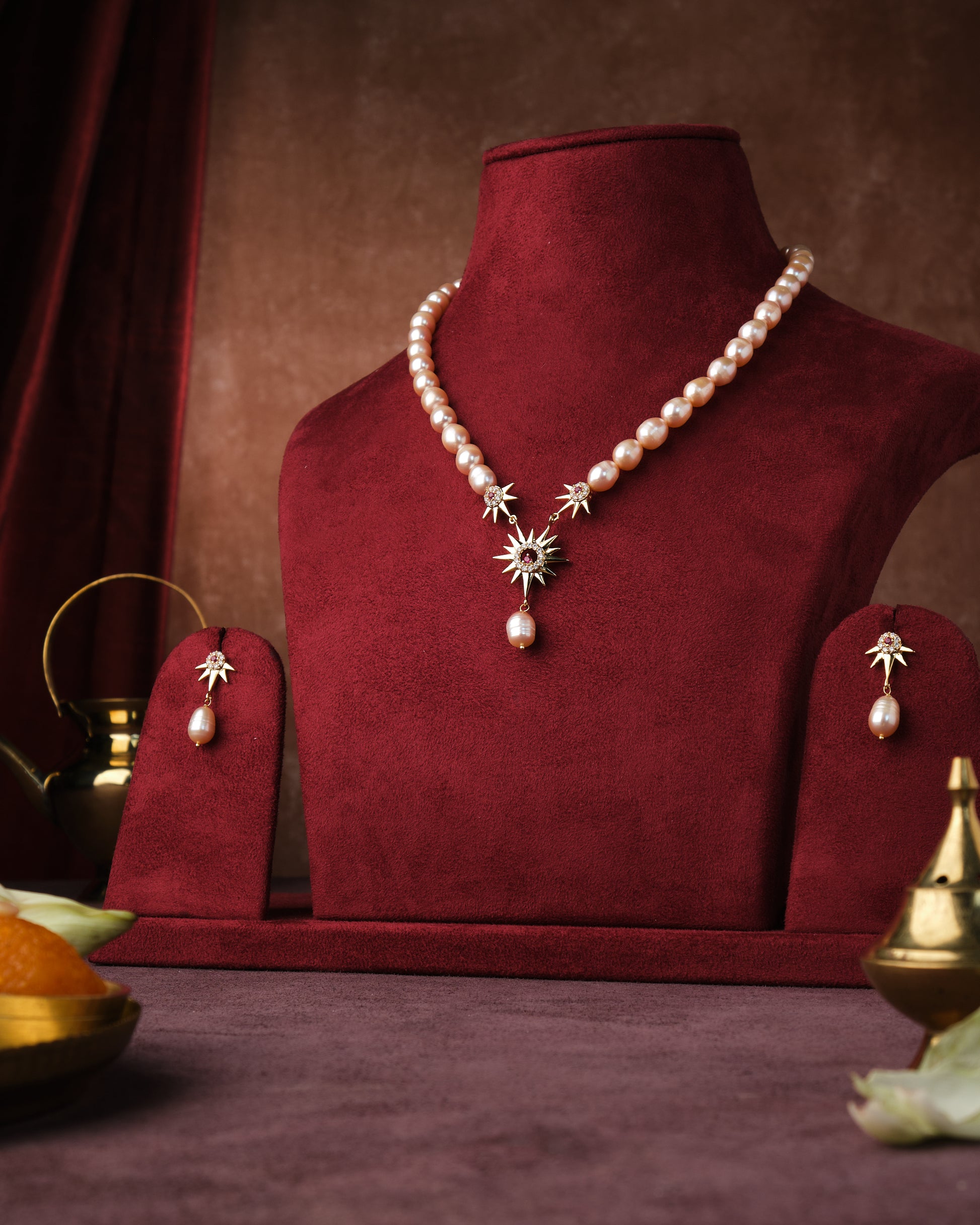 A Trendy Real Pearl Necklace Set by Chandrani Pearls India, featuring a sunburst pendant and matching earrings, displayed on a red velvet stand with decorative items in the background.